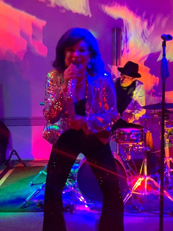 Ana singing on stage in a sparkling jacket under colorful lights, with the drummer behind her