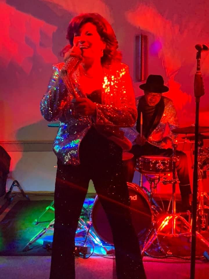 Ana singing in a sparkling jacket under red stage lights, with the drummer behind her