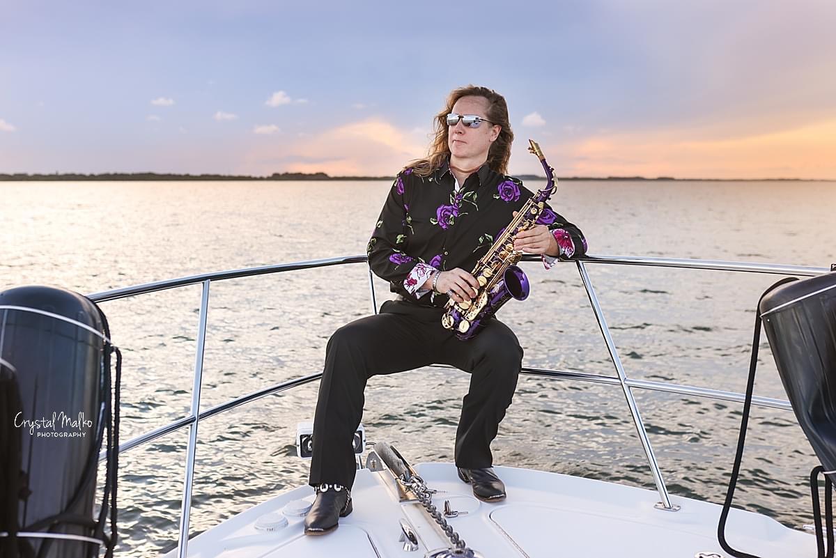 SWITCH saxophonist seated on a boat holding a purple saxophone at sunset